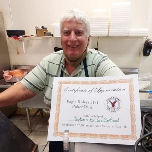 A smiling man in an apron holds a Certificate of Appreciation for Captain Brian's Seafood's donation.