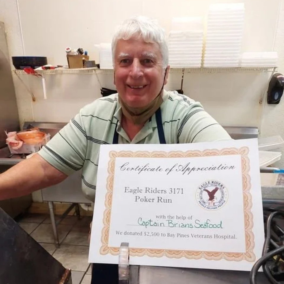 A smiling man in an apron holds a Certificate of Appreciation for Captain Brian's Seafood's donation.