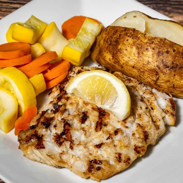 Grilled fish with a lemon slice, baked potato, and mixed vegetables on a white plate.